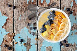 Pineapple, mango smoothie bowl with coconut, bananas, blueberries and granola, overhead view on wood