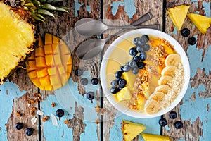 Pineapple, mango smoothie bowl with coconut, bananas, blueberries and granola, top view table scene on wood