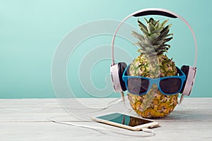 Pineapple with sunglasses, headphones and smart phone on wooden table over mint background. Tropical summer vacation