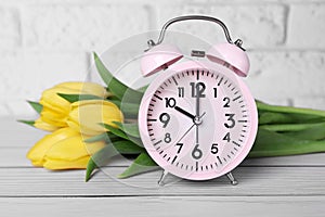 Pink alarm clock and beautiful tulips on white wooden table against brick wall, closeup. Spring time