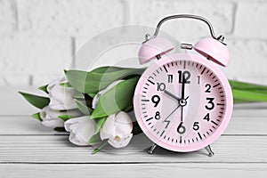 Pink alarm clock and beautiful tulips on white wooden table against brick wall, closeup. Spring time