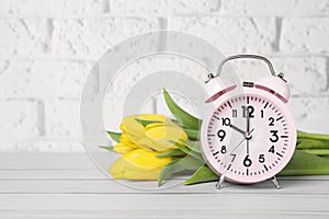 Pink alarm clock and beautiful tulips on white wooden table against brick wall, space for text. Spring time