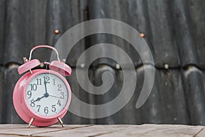 Pink alarm clock rests on an old wooden balcony and blurred background of gray tiled roof. classic style alarm clock is placed on