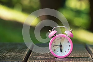 A pink alarm clock stands on a wooden board in the summer in the parks in the sun