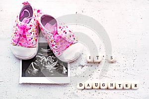 Pink baby booties with a picture of ultrasound for 20 weeks on a light background. Inscription it`s daughter.