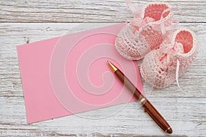 Pink baby booties with pink greeting card with pen on weathered wood