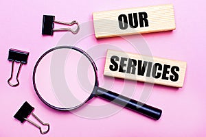 On a pink background, a magnifier, black paper clips and wooden blocks with the text OUR SERVICES. Business concept