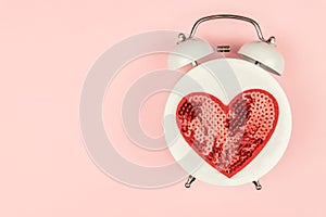 Pink Background with white alarm clock with red sequin heart. Creative layout. Top view. Time for love and greetings