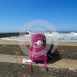 Pink backpack with scallop shell symbol of pilgrim at concrete wall with ocean with huge white waves and blue sky on background