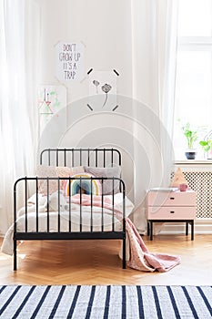 Pink blanket on bed next to cabinet in bright kid`s bedroom interior with posters and carpet