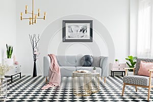 Pink blanket on grey sofa in living room interior with poster and patterned armchair. Real photo