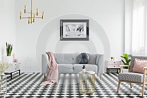 Pink blanket and pillows on grey couch in white living room interior with poster and armchair. Real photo