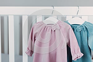 Pink, blue dresses for baby girl on white hanger near white bed in nursery. Set of baby clothes and accessories for