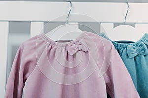 Pink, blue dresses for baby girl on white hanger near white bed in nursery. Set of baby clothes and accessories for