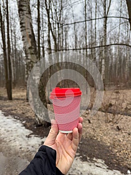 The pink cardboard coffee cup with a red plastic lid in men& x27;s hands on the background of a spring park, black jacket