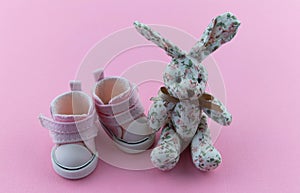 Pink children`s shoes on a delicate pink background. A textile rabbit is sitting nearby. Concept of first steps, birthday,