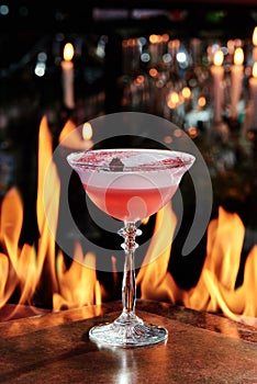 pink cocktail with white foam top in a beautiful glass on copper designer bar counter, the background of real fire