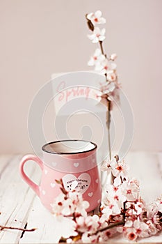 Pink coffee mug, white cherry flowers and `Spring` note; spring background