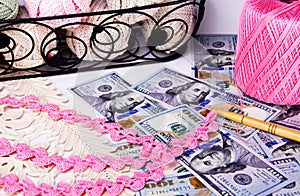 Pink crochet lace and crochet needle sitting on top of scattered hundred dollar bills and next to a basket filled with crochet