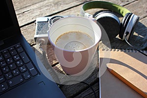 A pink cup of coffee, laptop, paper notebook, pencil, headphones, and vintage film camera on a wooden pier background.