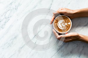 Pink cup of fresh cappuccino in woman hands on white marble table background