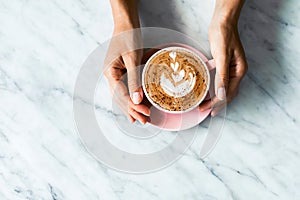 Pink cup of fresh cappuccino in woman hands on white marble table background