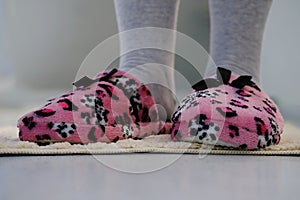 Pink domestic slipper with black bow on women`s legs, feet on a light plush rug, concept of comfortable shoes for rest, relax and