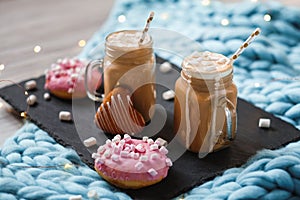 Pink donut with marshmallow and hot chocolate in glass cup on black tray on blue merino knit blanket. Lights on background