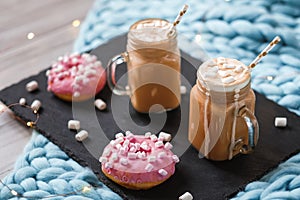 Pink donut with marshmallow and hot chocolate in glass cup on black tray on blue merino knit blanket. Lights on background