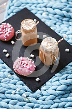 Pink donut with marshmallow and hot chocolate in glass cup on black tray on blue merino knit blanket. Lights on background