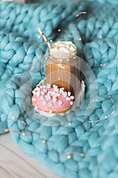 Pink donut with marshmallow and hot chocolate in glass cup on blue merino knit blanket. Lights on background