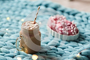 Pink donut with marshmallow and hot chocolate in glass cup on blue merino knit blanket. Lights on background