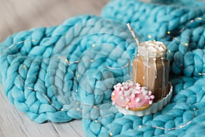 Pink donut with marshmallow and hot chocolate in glass cup on blue merino knit blanket. Lights on background