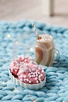 Pink donut with marshmallow and hot chocolate in glass cup on blue merino knit blanket. Lights on background