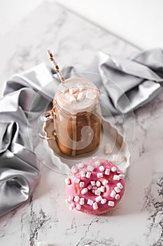 Pink donut with marshmallow and hot chocolate in glass cup on marble table.