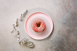 Pink donut on plate, measuring tape over grey concrete background. Diet concept. Weight lost after holidays