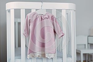 Pink dress for baby girl on white hanger near white bed in nursery. Set of baby clothes and accessories for birthday