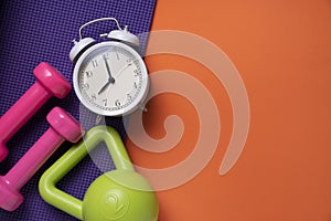 Pink dumbbell, alarm clock, kettle bell and purple yoga mat on orange table background