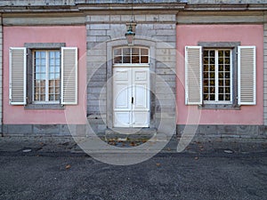 The pink facade of the historic building with white old wooden entrance doors and windows. There are wooden shutters in the window