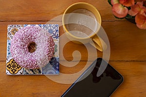 Pink frosted donut on the wooden table, coffee cup and cellphone - pause or breakfast
