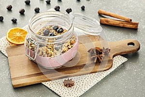 Pink, fruit yogurt with granola and blueberries in a jar on a wooden cutting board and cinnamon spice on a gray kitchen counter