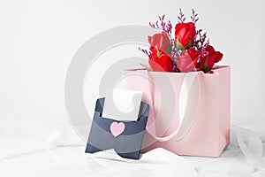 Pink gift bag with rose and card in black envelope with heart on white table with ribbon