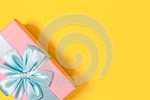 Pink gift box tied with blue ribbon with bow at the top on yellow background. Copy space for text. Minimal flat lay. Top view.