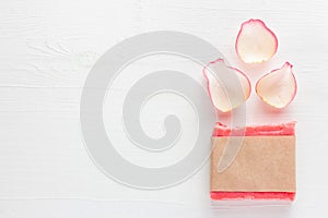 Pink handmade floral soap bar in blank paper label package next to rose petals on white table background