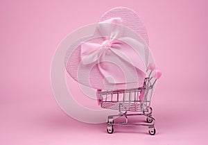 Pink heart gift with bow for Valentine`s Day in shopping scart on a pink background, delivery gift