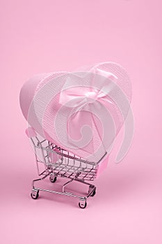 Pink heart gift with bow for Valentine`s Day in shopping scart on a pink background, minimalism