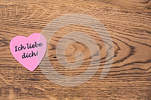 Pink heart post-it with the text `Ich liebe dich` on a wooden background. Translation: `I love you`