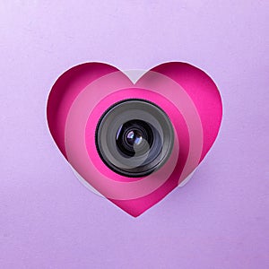 Pink heart on a purple background. At the heart is a camera lens. Concept of love, photography and video
