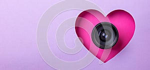 Pink heart on a purple background. At the heart is a camera lens. Concept of love, photography and video. Copy space