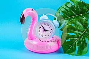 Pink inflatable flamingo on blue background with monstera leaves and clock.